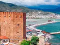 Alanya zwiedzanie na własną rękę: potężna czerwona wieża zamku, port z łodziami, latarnia morska i miasto w tle.