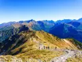 Grupa turystów wędruje granią górską, podziwiając rozległą panoramę. W oddali widać kolejne szczyty, tworzące zapierającą dech w piersiach przepaść w górach.