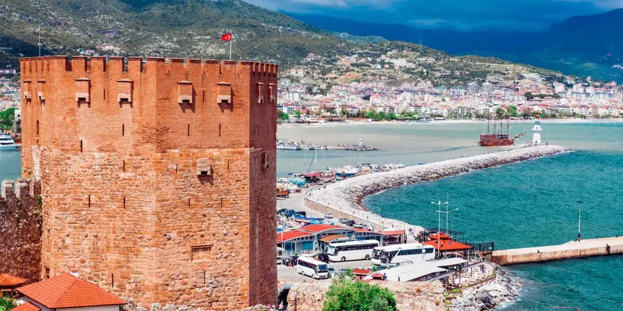 Alanya zwiedzanie na własną rękę: potężna czerwona wieża zamku, port z łodziami, latarnia morska i miasto w tle.
