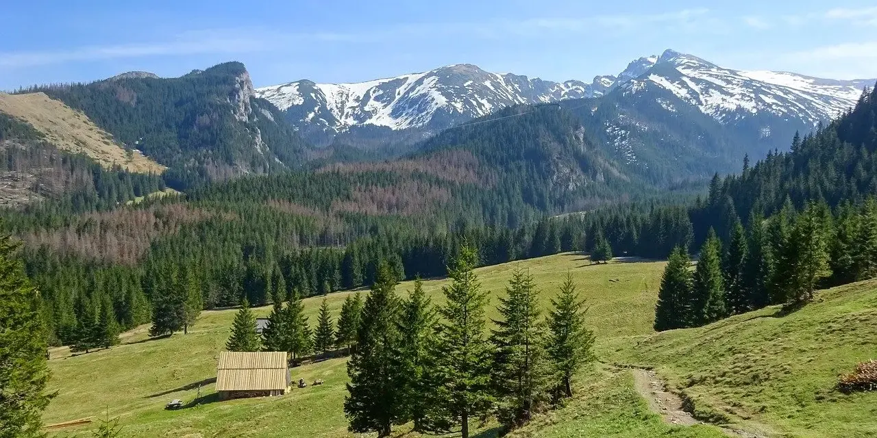 Piękne jezioro w górach, otoczone lasem i ośnieżonymi szczytami. W oddali widać drewniany domek.