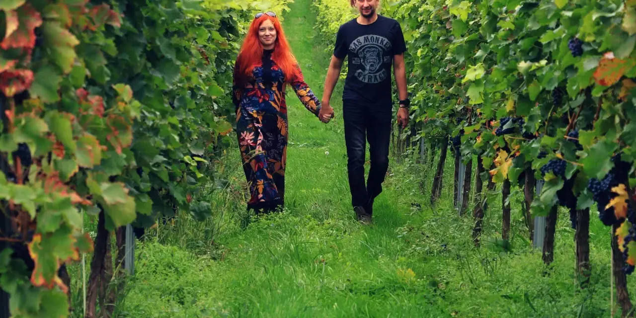Para spaceruje po winnicy na Dolnym Śląsku, trzymając się za ręce. Wokół bujne zielone liście i dojrzałe winogrona.