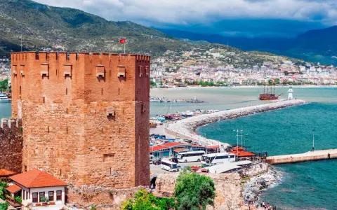 Alanya zwiedzanie na własną rękę: potężna czerwona wieża zamku, port z łodziami, latarnia morska i miasto w tle.