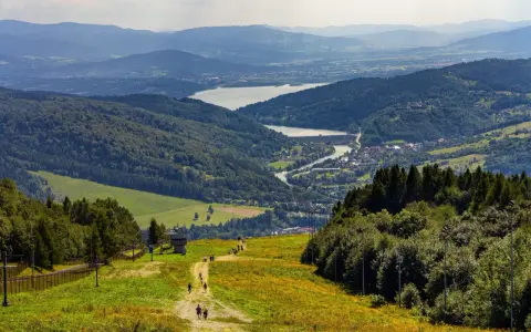 Widok na góry, jezioro i miasteczko. Turyści idą ścieżką, myśląc, gdzie w góry pociągiem wybrać się na wycieczkę.