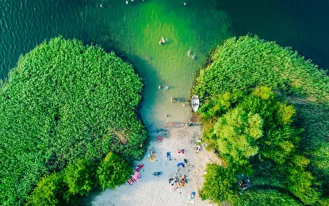 Letnie orzeźwienie nad jednym z najpiękniejszych jezior na południu Polski. Ludzie kąpią się, plażują, a łódka czeka przy brzegu.