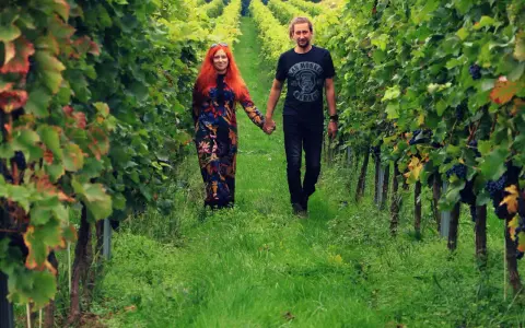 Para spaceruje po winnicy na Dolnym Śląsku, trzymając się za ręce. Wokół bujne zielone liście i dojrzałe winogrona.