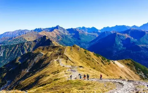 Grupa turystów wędruje granią górską, podziwiając rozległą panoramę. W oddali widać kolejne szczyty, tworzące zapierającą dech w piersiach przepaść w górach.