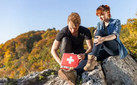 Mężczyzna z czerwoną apteczką pomaga kobiecie po wypadku w górach. Jesienne drzewa w tle.