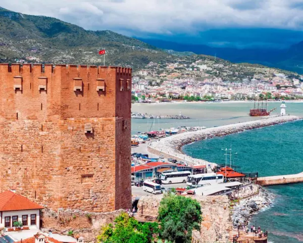 Alanya zwiedzanie na własną rękę: potężna czerwona wieża zamku, port z łodziami, latarnia morska i miasto w tle.