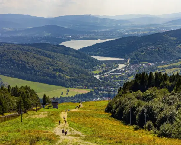 Widok na góry, jezioro i miasteczko. Turyści idą ścieżką, myśląc, gdzie w góry pociągiem wybrać się na wycieczkę.