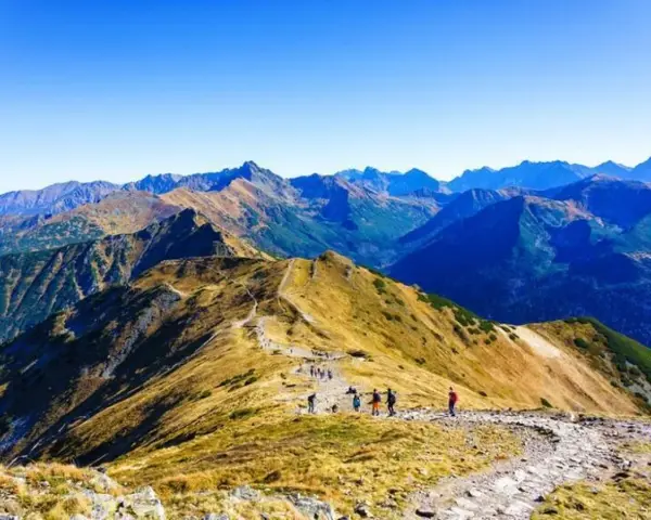 Grupa turystów wędruje granią górską, podziwiając rozległą panoramę. W oddali widać kolejne szczyty, tworzące zapierającą dech w piersiach przepaść w górach.