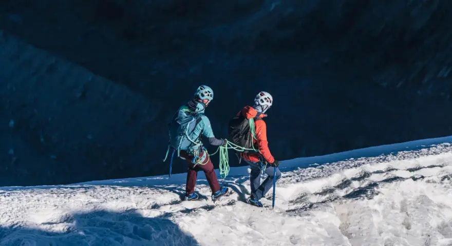 Dwóch alpinistów z rakami i czekanami wspina się po ośnieżonym zboczu.