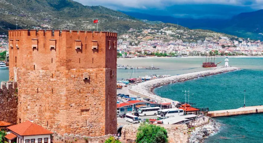 Alanya zwiedzanie na własną rękę: potężna czerwona wieża zamku, port z łodziami, latarnia morska i miasto w tle.