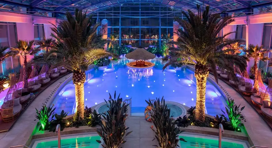 Wnętrze hotelu z basenami termalnymi, palmami i podświetlanym barem w wodzie. Relaks w tropikalnym klimacie.