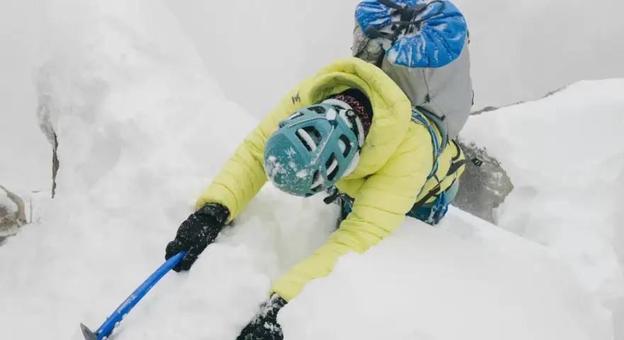 Wspinaczka w śniegu, pokazująca jak trzymać czekan. Osoba w żółtej kurtce i kasku wspina się po oblodzonej skale.