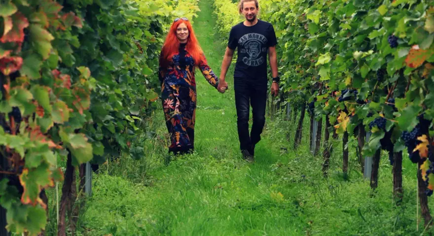 Para spaceruje po winnicy na Dolnym Śląsku, trzymając się za ręce. Wokół bujne zielone liście i dojrzałe winogrona.