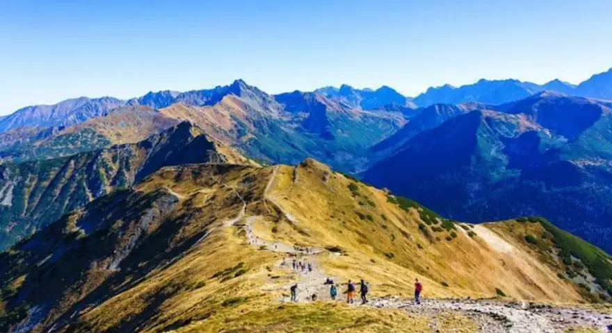 Grupa turystów wędruje granią górską, podziwiając rozległą panoramę. W oddali widać kolejne szczyty, tworzące zapierającą dech w piersiach przepaść w górach.