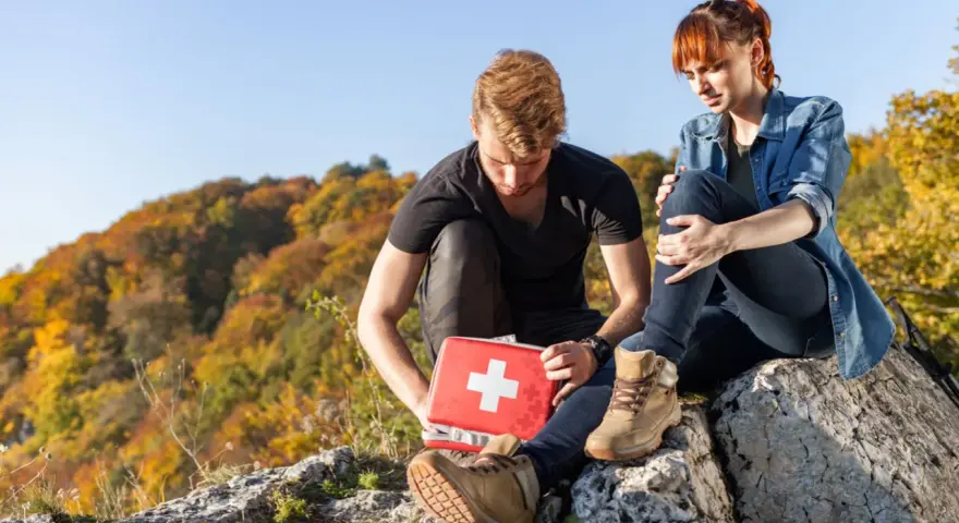 Mężczyzna z czerwoną apteczką pomaga kobiecie po wypadku w górach. Jesienne drzewa w tle.