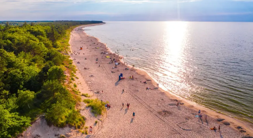 Piękna plaża nad Bałtykiem, idealna na fajne i tanie miejsca na wakacje w Polsce. Słońce odbija się w wodzie, a las tworzy malownicze tło.