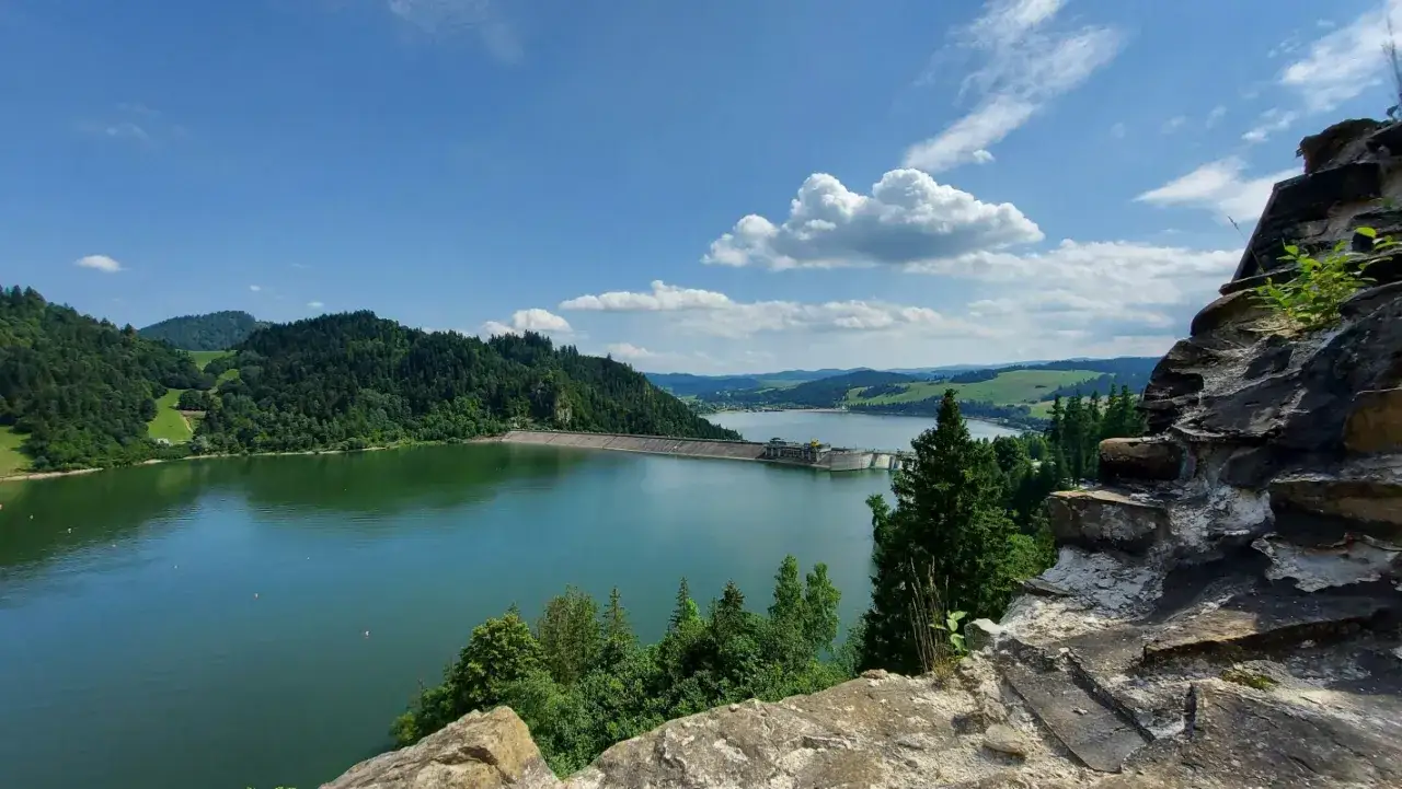 Malownicze jezioro w górach otoczone zielonymi lasami, zaporą wodną i skalistym zboczem w pierwszym planie.