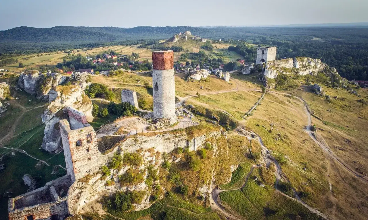 Ruiny zamków na wapiennych skałach, w tym okrągła wieża, tworzą malowniczy krajobraz. W oddali widać wieś na szlaku Orlich Gniazd.