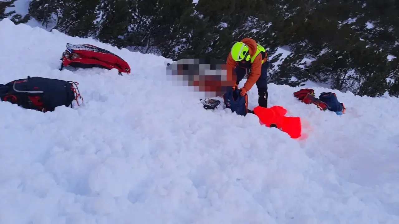Akcja ratunkowa po wypadku w g&oacute;rach. Ratownik w pomarańczowej kurtce i kasku pomaga przy osobie poszkodowanej w śniegu. Wok&oacute;ł leżą plecaki i sprzęt.