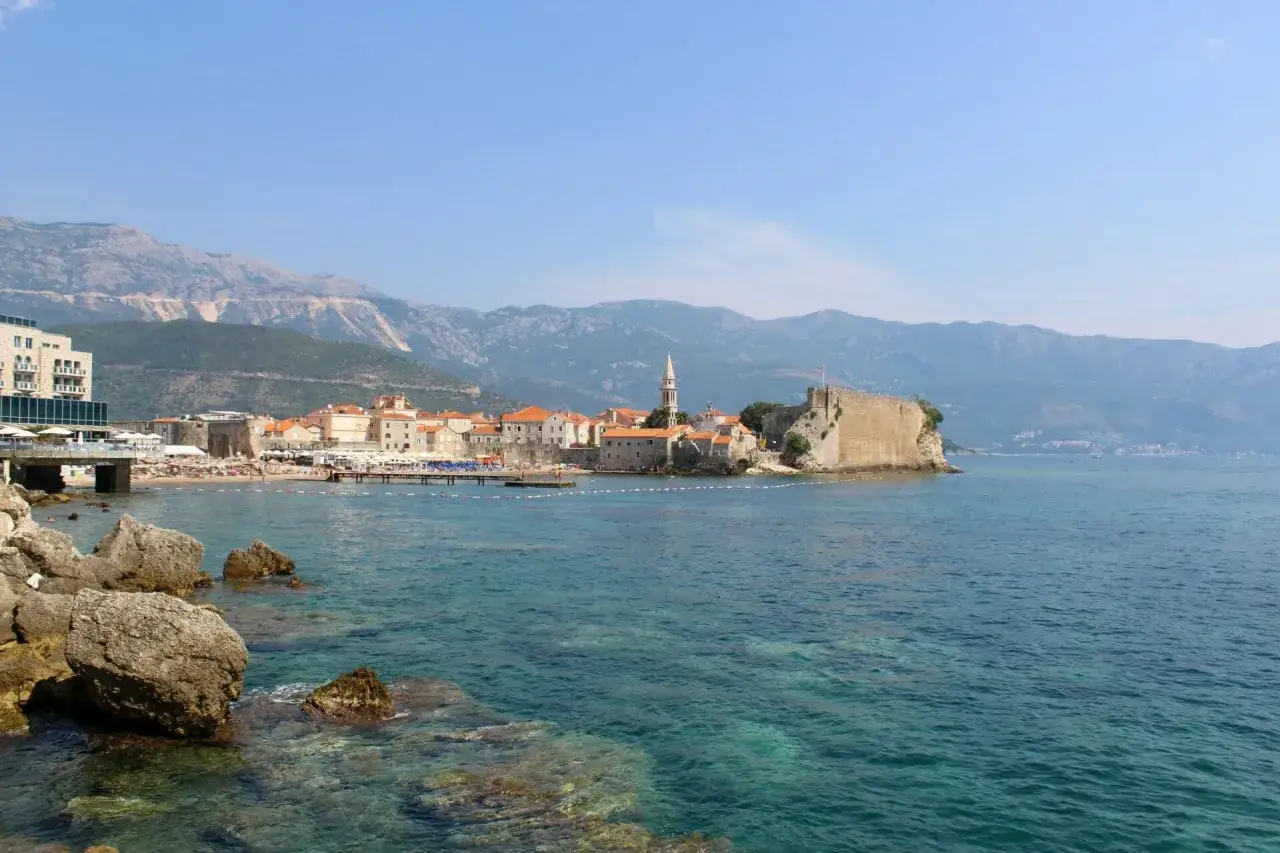 Widok na budva stare miasto z twierdzą i kościołem na skalistym cyplu, otoczone turkusowym morzem i górami w tle.